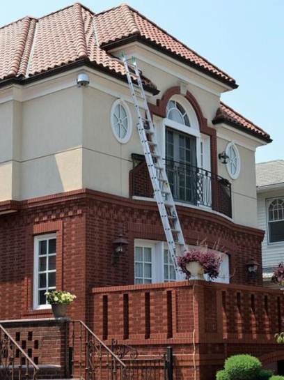 A tall ladder reaches to the top of a large home where roof maintenance workers make repairs.